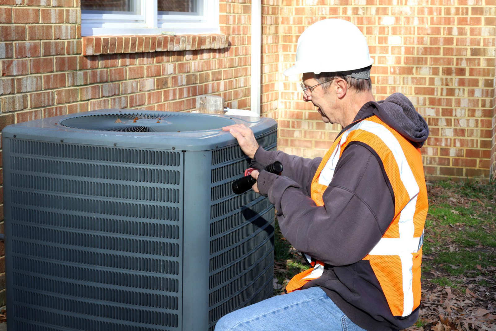 A professional HVAC technician wearing a hard hat and safety vest using a flashlight to inspect an outdoor air conditioner unit for repairs.