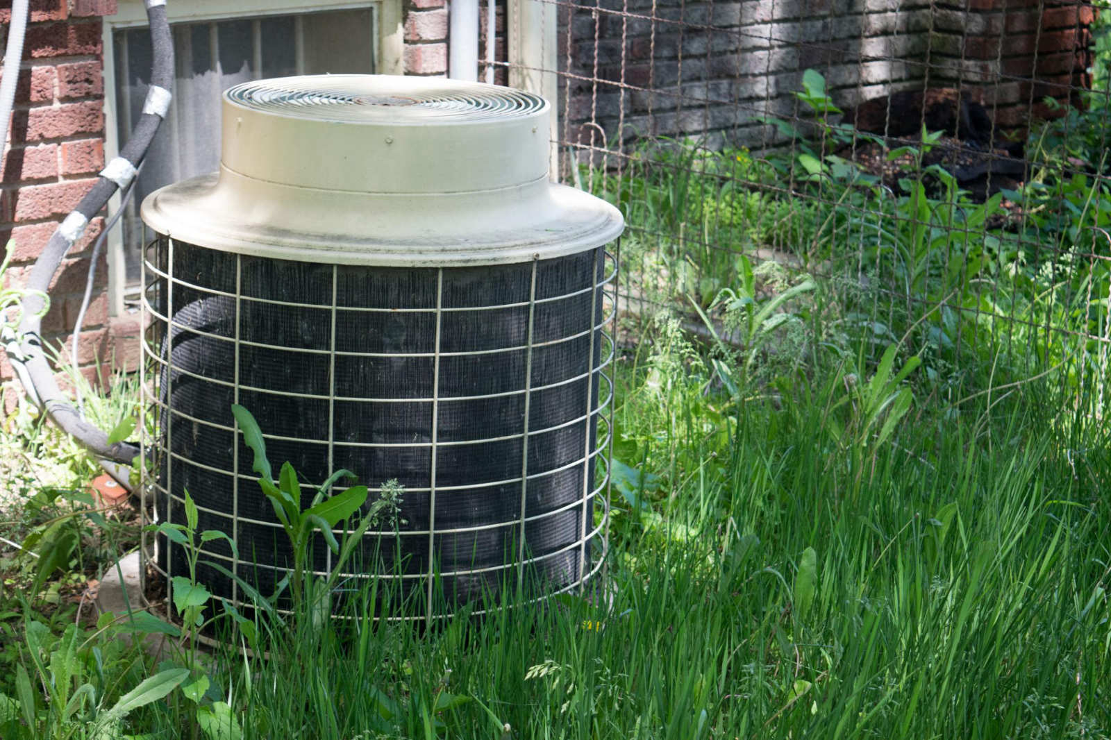 An aging, weathered air conditioning unit sitting in tall grass outside a brick home, illustrating a system nearing the end of its lifespan.