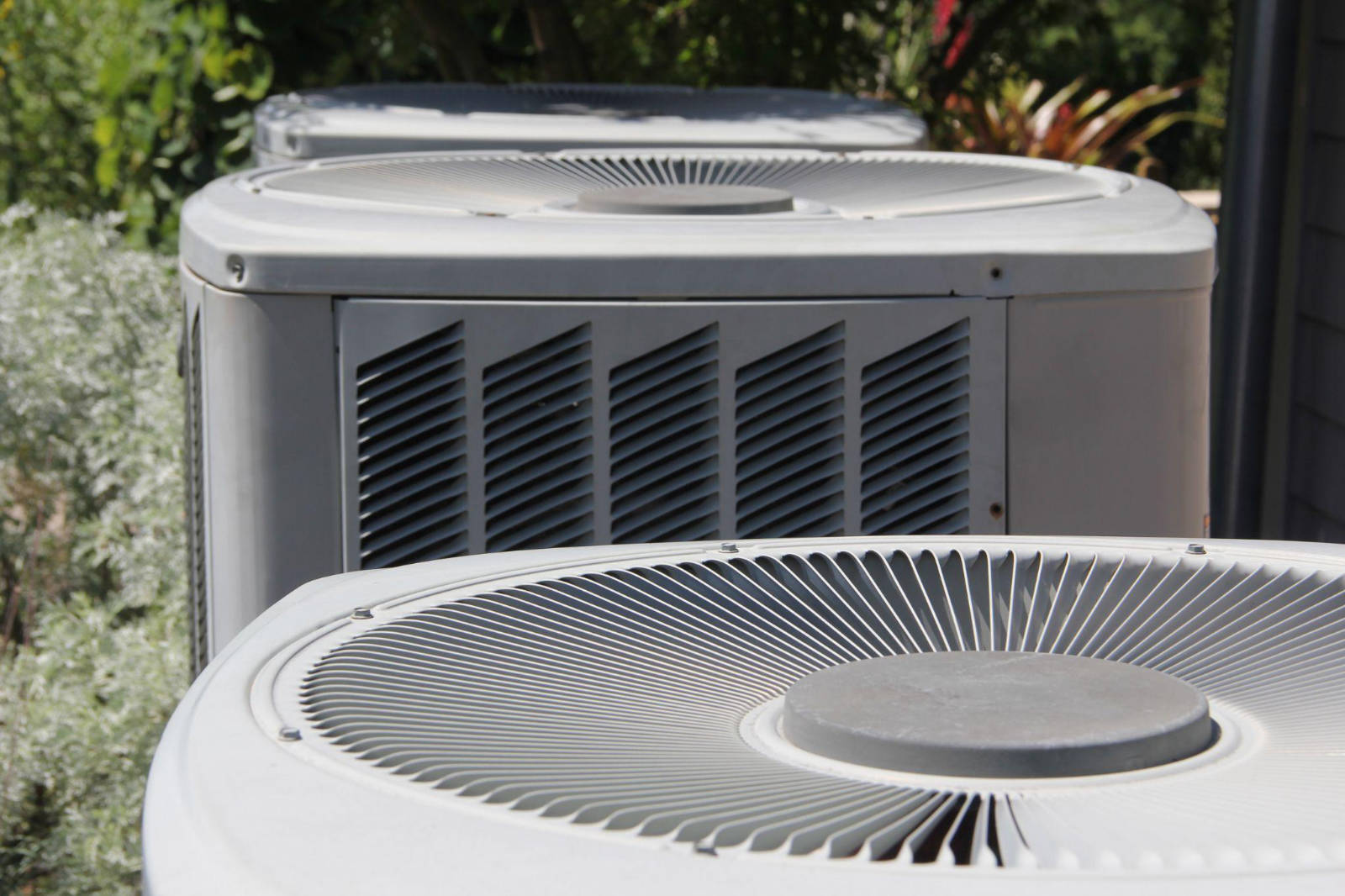 A row of outdoor air conditioner units sitting next to garden vegetation requiring regular clearing and maintenance.