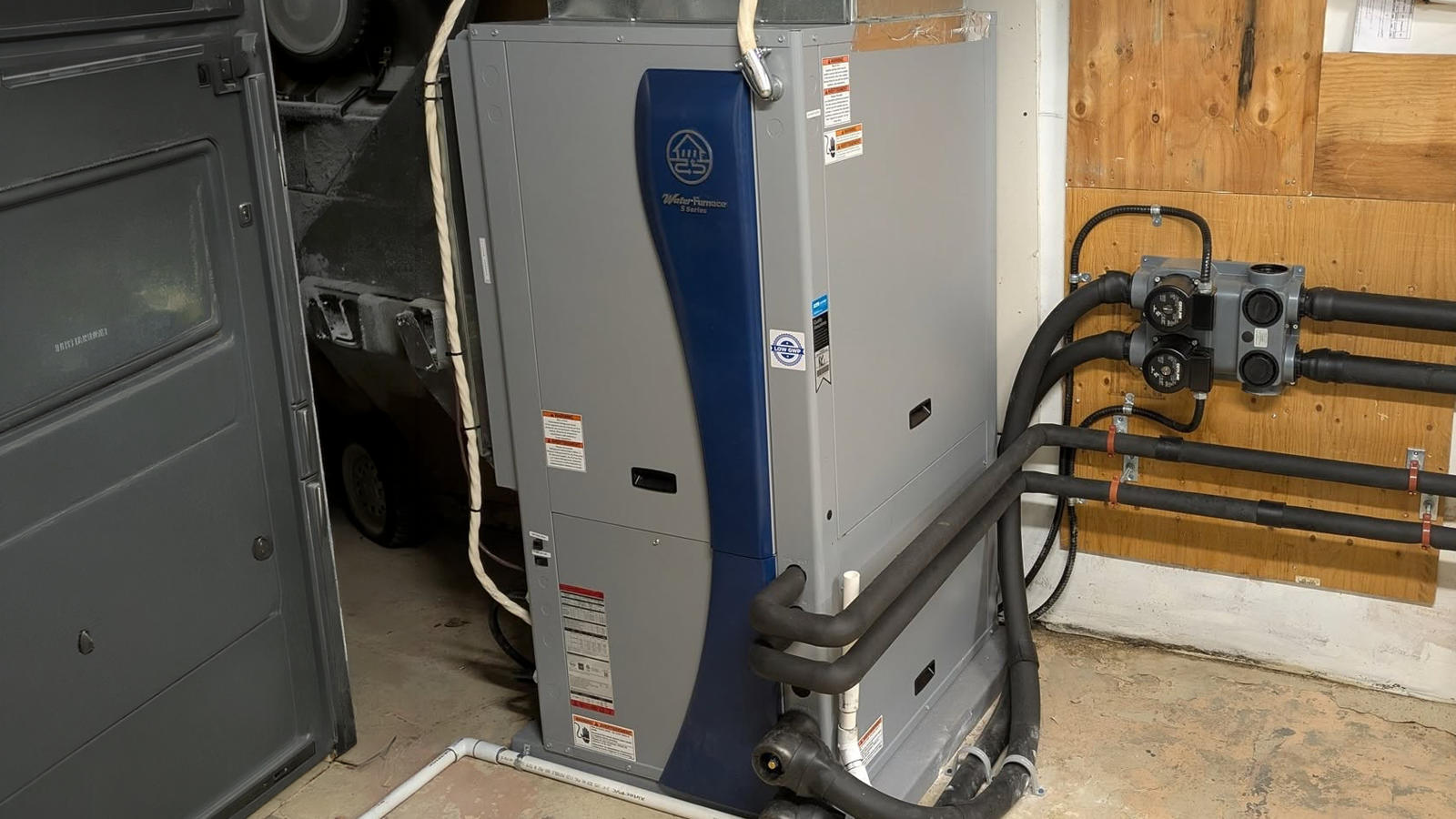 A gray and blue WaterFurnace heating unit with insulated piping installed in a home basement.